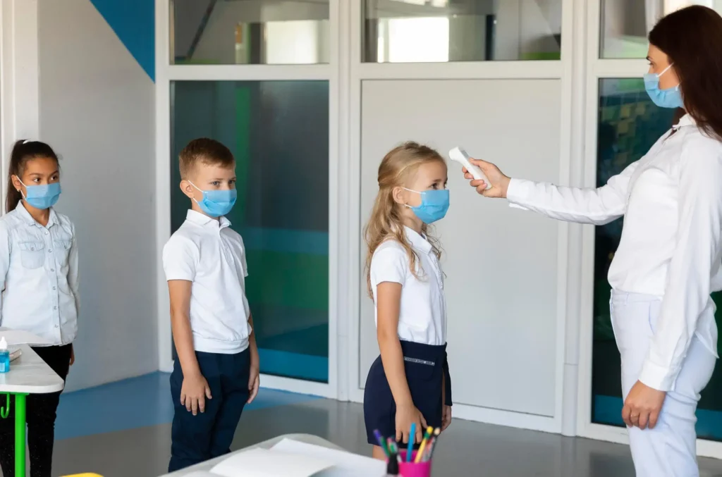 A healthcare provider in a face mask uses a thermometer to check a student's temperature in a medical evaluation room 