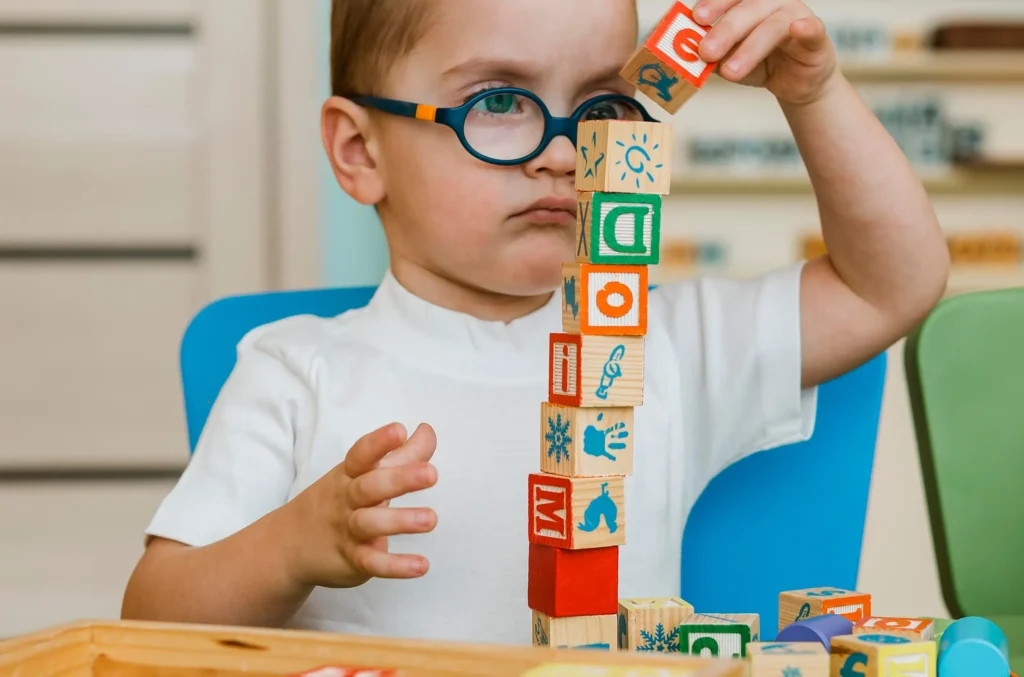 A young child wearing glasses is focused on building with wooden blocks, which forms a crucial part of developmental and speech evaluation
