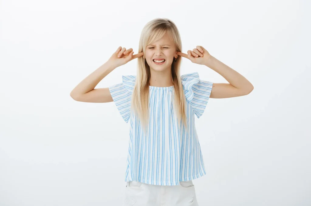 A young girl wearing a blue and white striped shirt is covering her ears, indicating discomfort related to noise or the need for earwax removal and cleaning.