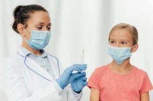 A doctor wearing a mask and gloves administers a vaccine to a calm young girl in medical setting