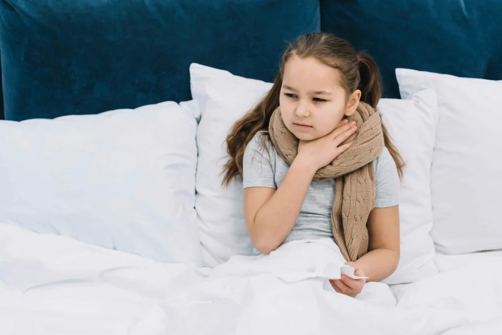 A young girl sitting on a bed, wrapped in a blanket, suffering from a sore throat