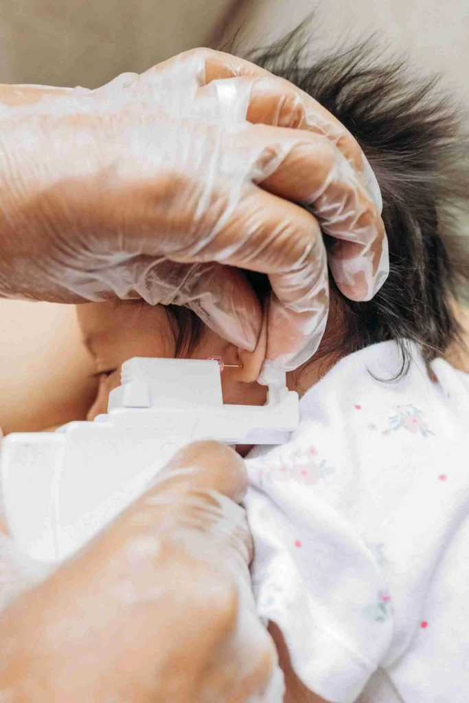 A caregiver wearing medical gloves gently holds a calm baby in a sterile environment, preparing for ear piercing with a small earring visible in the foreground.