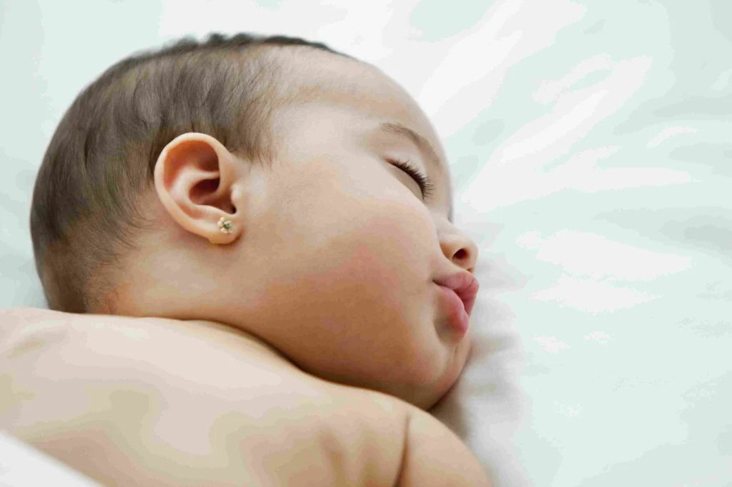 A close-up of a baby peacefully sleeping on a cozy bed, with freshly pierced ears visible.