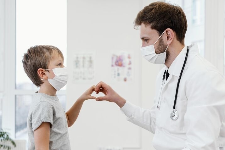 A masked young boy grips a doctor's hand, representing trust and reassurance during a healthcare visit.