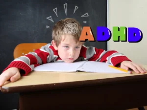 A tired boy sitting at a desk, resting his head on a notebook, with 'ADHD' in colorful letters above him.