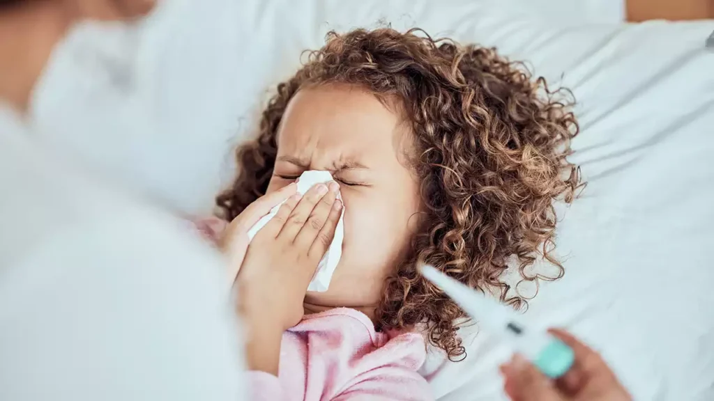 A sick child with flu symptoms in New Jersey, sneezing into a tissue while a caregiver checks her temperature with a thermometer.
